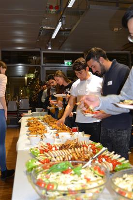 A Long table is filled with salads and different finger Food dishes. International students are Standing in line along the table and filling their plates with Food.